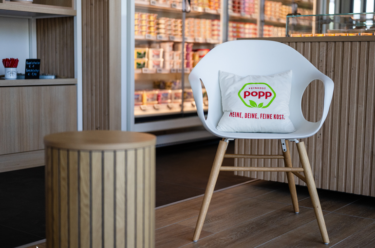 Ein Stuhl, der in einem Raum steht. Nebenan ist ein kleiner Tisch. Auf dem Stuhl liegt ein Kissen mit einem Popp Logo.